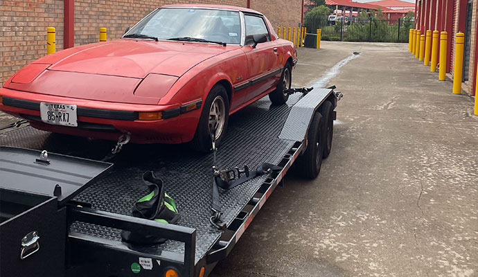 Red classic Mazda RX-7 secured on a black PJ car hauler trailer with straps.