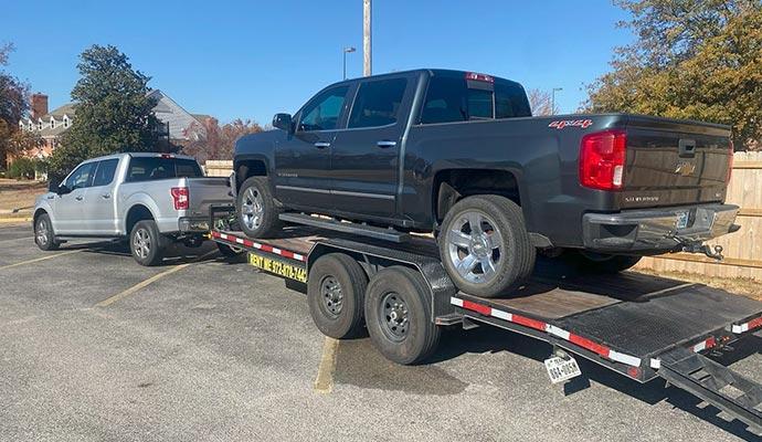 An 83 x 20 PJ car hauler trailer carrying a black pickup truck, towed by a silver pickup truck