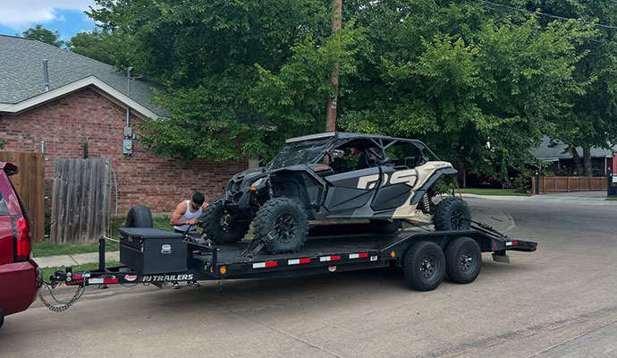 Person kneeling beside a Can-Am Maverick R side-by-side vehicle on a black flatbed trailer