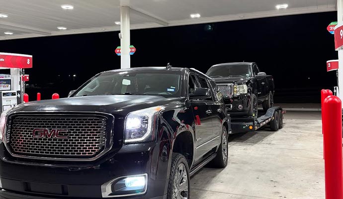Car hauling another black car on a trailer at a gas station
